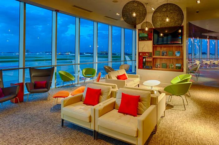 The lounge features comfortable beige-upholstered chairs with red pillows, green modern curved seating, and a wall of windows that overlook the airport runways.