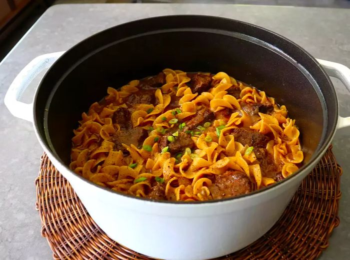 A white Dutch oven with beef and curly egg noodles displayed on a rattan mat, placed on a gray countertop.