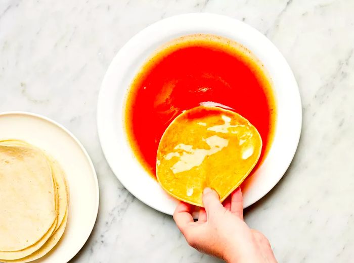 Hand dipping a corn tortilla into red enchilada sauce on a white plate