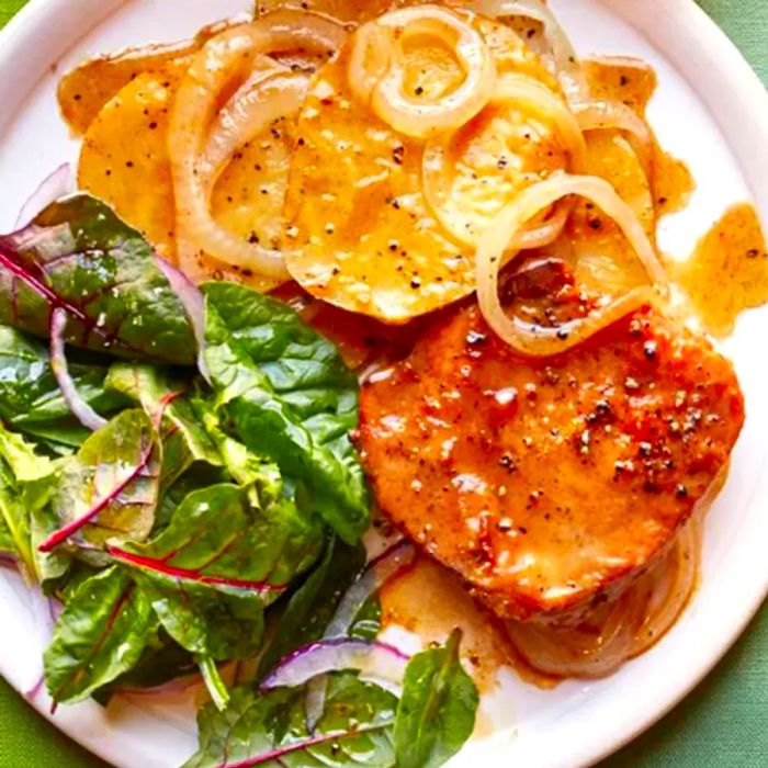 A close-up view of skillet-cooked pork chops with potatoes and onions, served alongside a fresh salad on a white plate.