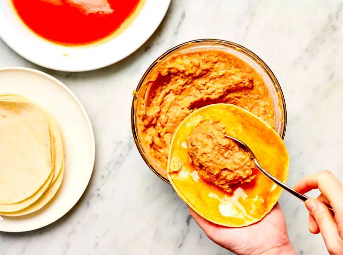 Hand spooning refried beans onto a corn tortilla topped with red enchilada sauce