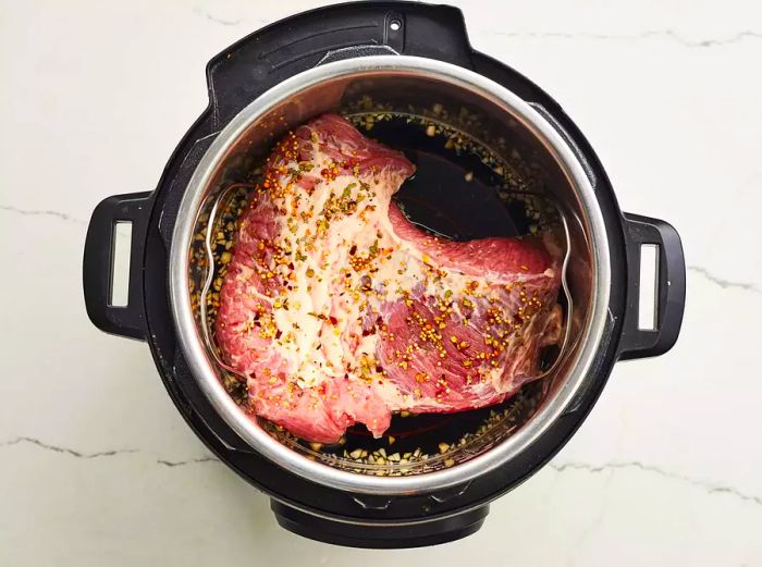 A top-down view of a pressure cooker with a raw beef brisket resting inside, topped with spices.