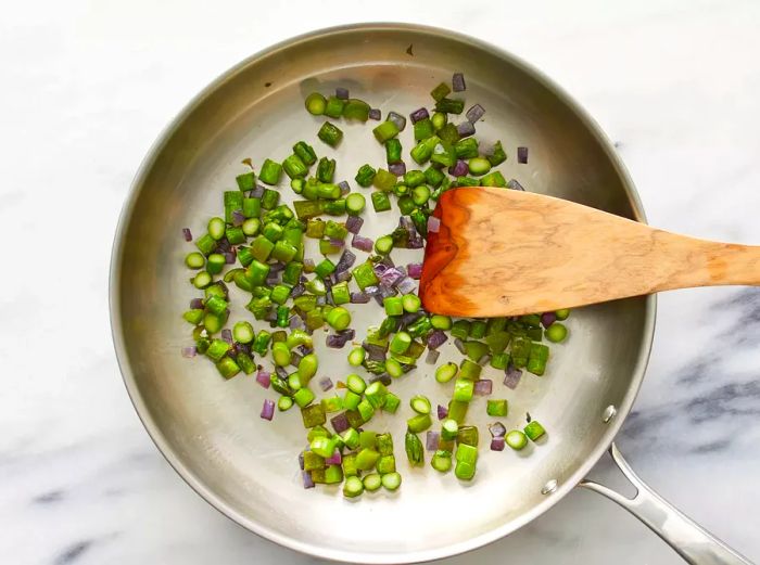 A top-down view of asparagus, green bell pepper, and onion sizzling in a skillet, stirred with a wooden spoon.