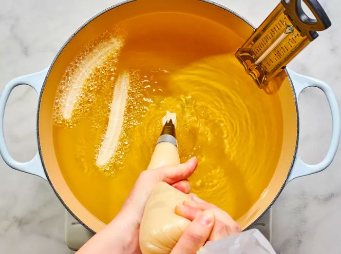 Two hands piping dough into a large pot of sizzling oil.