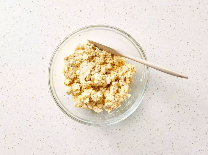 A glass bowl containing ricotta, walnuts, Parmesan cheese, breadcrumbs, and oregano, stirred with a wooden spoon.