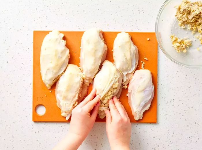 A cutting board with five chicken breasts with skin on, as two hands stuff the ricotta mixture beneath the skin of one breast.