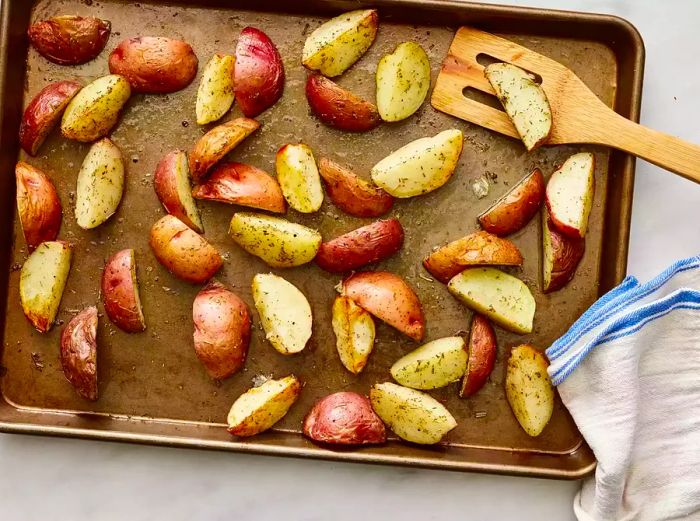 Baked, seasoned potatoes on a tray with a dish towel and wooden spoon placed to the side.