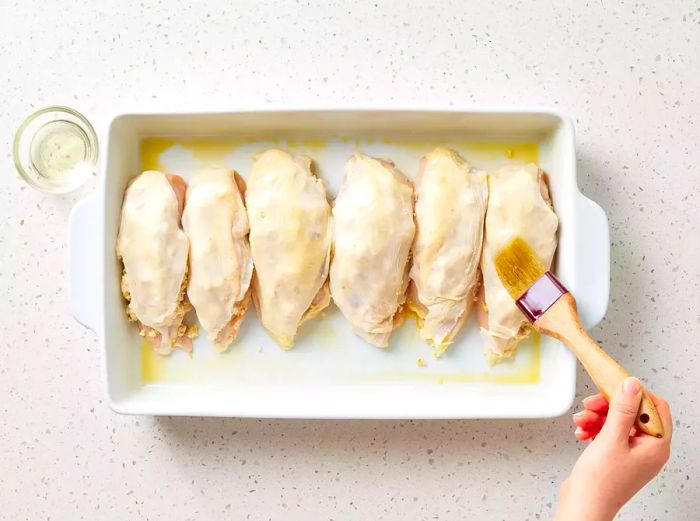 A baking dish filled with stuffed chicken breasts, being brushed with oil.