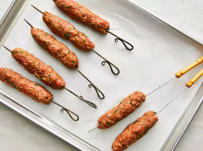 High-angle view of kofta kebabs being assembled on skewers, placed on a baking sheet.