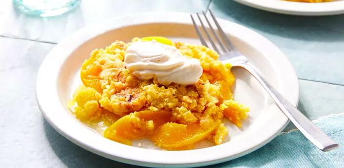 A serving of peach cobbler dump cake on a white plate, topped with a dollop of whipped cream and a silver fork beside it.