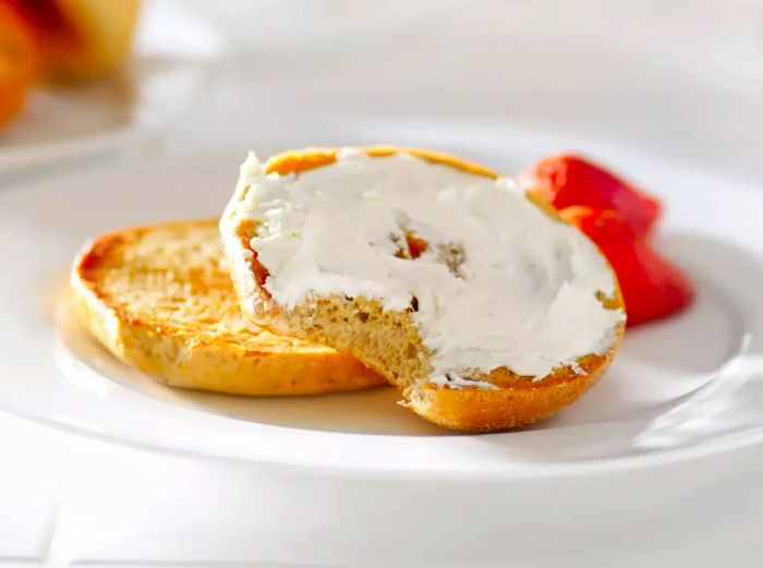 A toasted bagel, sliced and stacked with cream cheese spread on the top half, placed on a plate next to fresh strawberries.