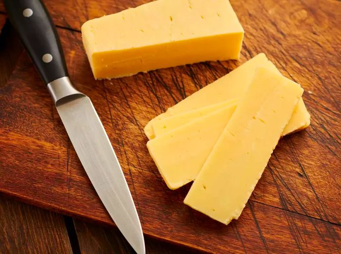 A block of white cheddar and slices on a wooden cutting board with a knife beside it.