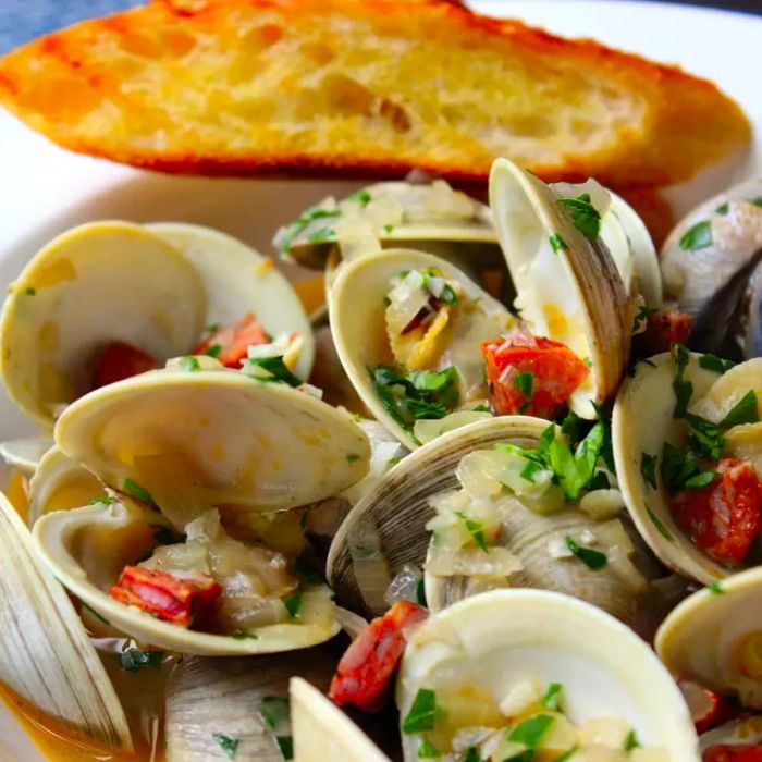 A close-up of Chorizo Steamed Clams with onions, fresh herbs, and a slice of toast, served in a white bowl