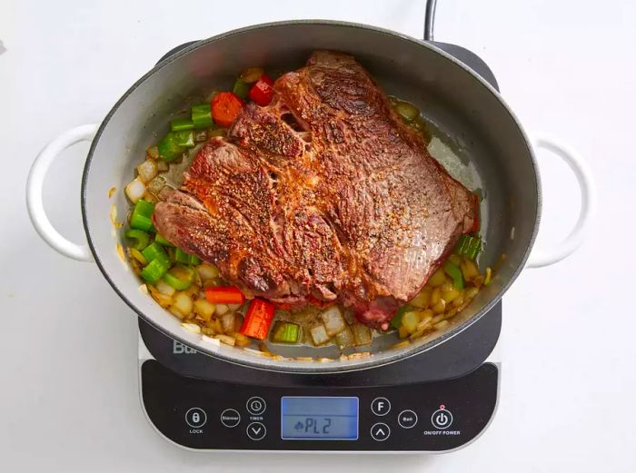 A large pot featuring a golden-brown chuck roast resting on a bed of chopped vegetables.