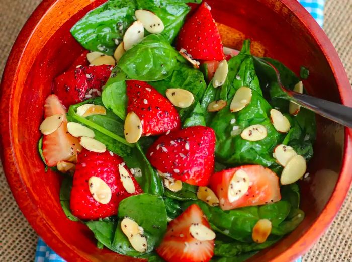 a top-down view of a wooden bowl filled with spinach salad, topped with strawberry halves, sliced almonds, and a poppyseed dressing