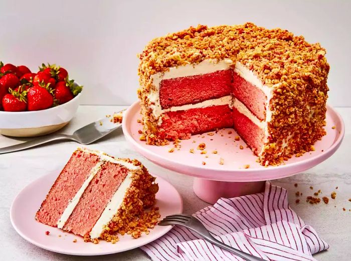 A large slice of strawberry crunch cake on a plate, with the remaining cake sitting on a cake stand in the background.