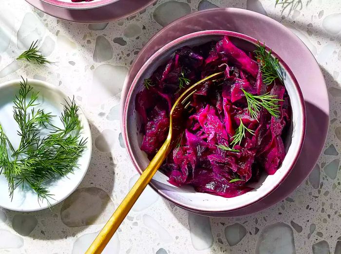 An overhead shot of Grandma Jeanette's Delicious German Red Cabbage, topped with fresh dill, served in a bowl with a gold fork beside it, accompanied by more fresh dill on a white plate.