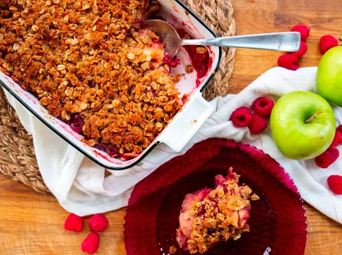 A close-up of a scoop of Apple-Raspberry Crisp on a plate, with another scoop still in a baking dish surrounded by fresh raspberries and apples.