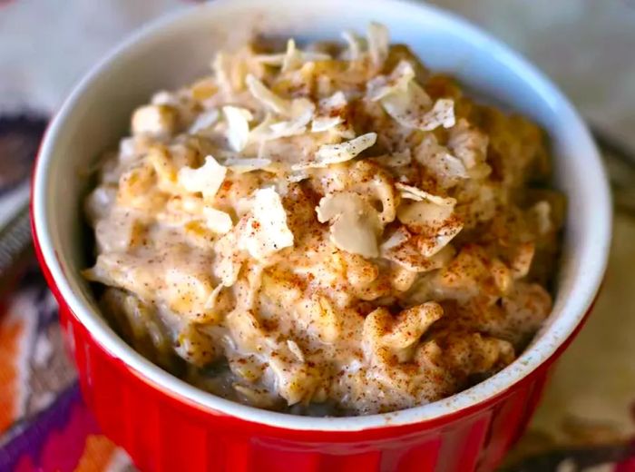 A close-up shot of sweet coconut oatmeal, topped with cinnamon and coconut, served in a red and white ramekin.