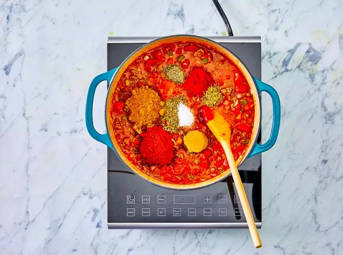 A top-down view of the chili mixture, with various seasonings and spices scattered on top, ready to be stirred in.