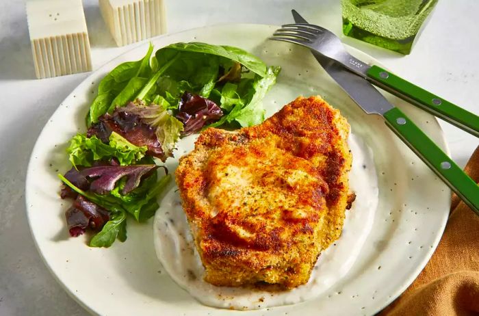 A high-angle view of a baked pork chop served on a rich mushroom cream sauce, accompanied by dressed mixed greens.