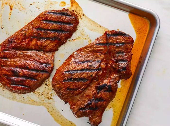 A steak resting on a baking sheet with marinade dripping beneath it.