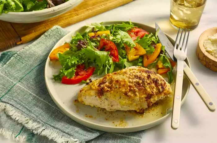 A single serving of mayo-coated chicken breast paired with a fresh green salad and ripe tomatoes.