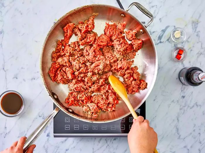 Ground beef cooking and being stirred in a skillet, with other ingredients set aside.