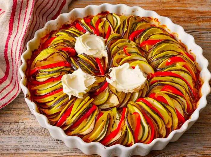 A close-up of a baking dish filled with Disney’s Ratatouille, topped with a few spoonfuls of mascarpone cheese.