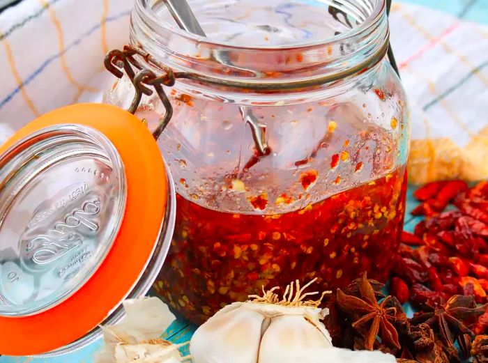 Close-up of homemade chili crisp in a jar, surrounded by garlic, star anise pods, and peppers.