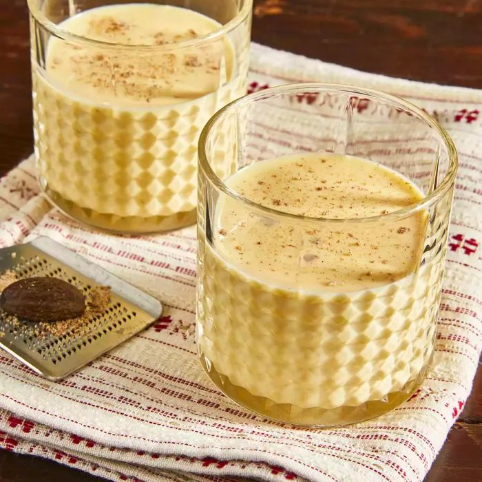 A close-up view of two glasses of irresistible eggnog, with fresh nutmeg and a grater nearby.