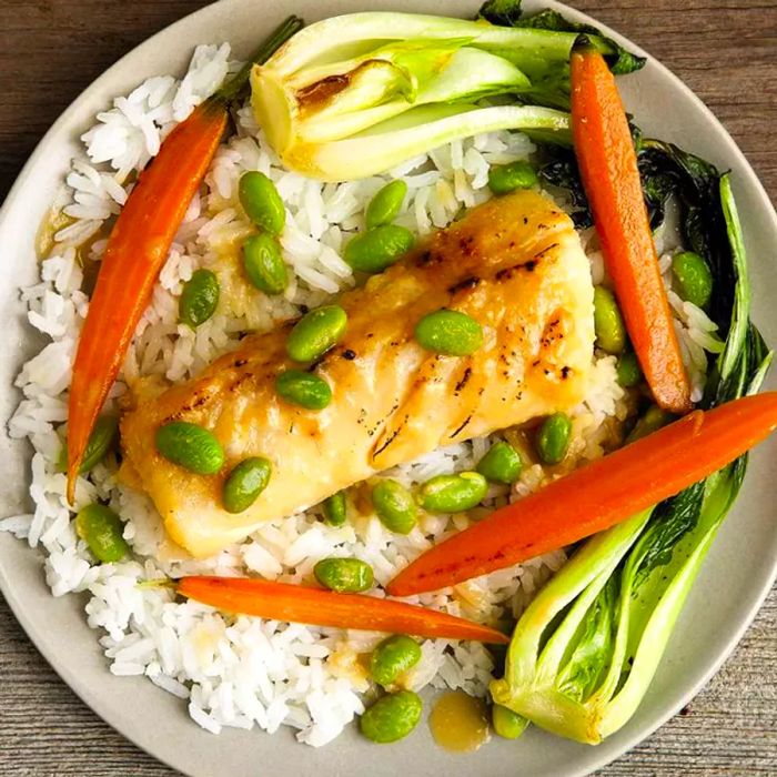 Close-up of a Miso-Glazed Black Cod fillet plated with white rice, carrots, bok choy, and edamame.