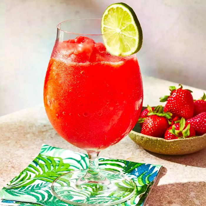 A frozen strawberry daiquiri, garnished with lime, rests on a table alongside a bowl of fresh strawberries and a tropical-themed linen.