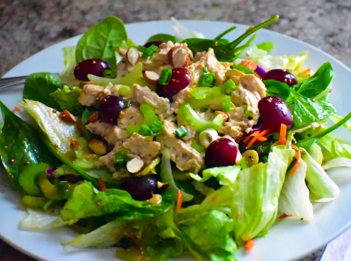 a mixed green salad topped with turkey salad, diced celery, halved red grapes, and scallions on a white plate