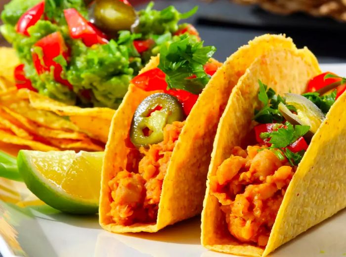 A close-up of Vegetarian Chickpea Tacos served alongside chips, guacamole, and fresh limes on a plate