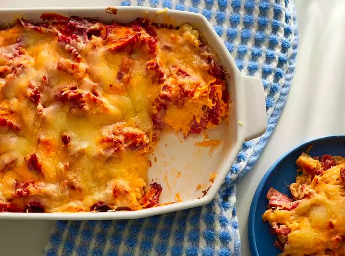 A close-up view of a Reuben casserole in a white baking dish with a slice served on a blue plate.