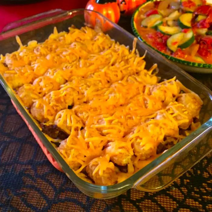 A rectangular glass baking dish filled with a cheesy Tater Tot topping over a ground beef casserole, with a vibrant side of zucchini and tomatoes in the background.