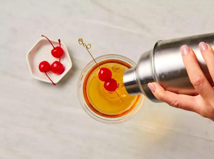 The cocktail is poured into the glass and topped with two maraschino cherries.