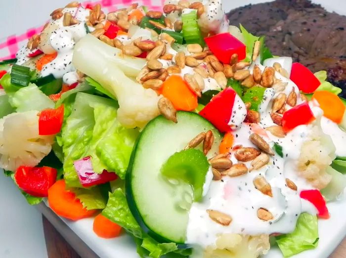 a colorful salad with romaine lettuce, cucumber, cauliflower, carrot, red bell pepper, and other fresh veggies, topped with a creamy dressing and roasted sunflower seeds