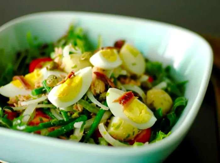 a white bowl filled with salad greens topped with tuna, cooked green beans, anchovy fillets, quartered hard-cooked eggs, and capers