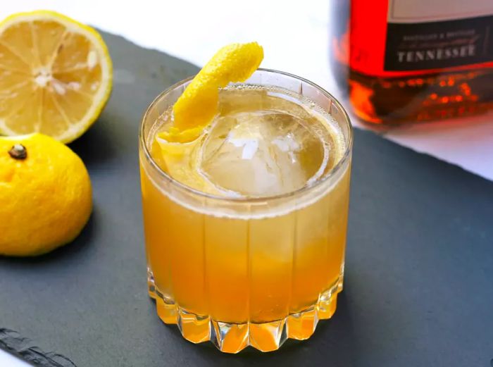 A golden cocktail served in a highball glass with fresh lemons and whiskey in the background