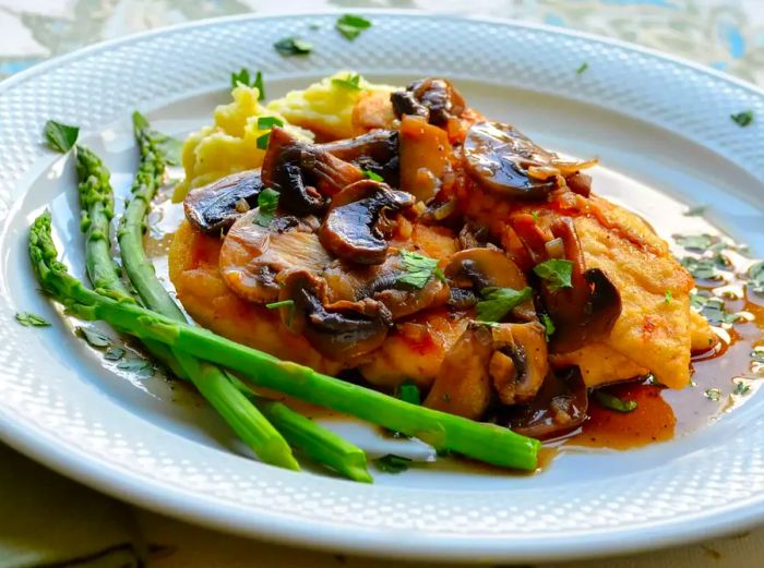 Close-up view of Chicken Madeira with mushrooms, served with asparagus and mashed potatoes, garnished with fresh herbs on a white plate.