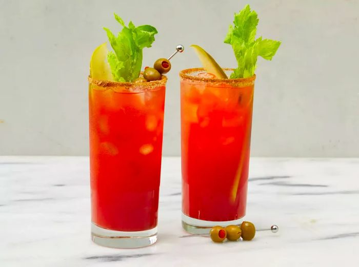 A mid-angle view of two highball glasses filled with Canadian Caesars, garnished with celery, pickle spears, and olives.