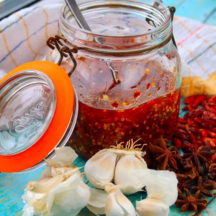 A jar of homemade chili crisp, made with dried chili flakes and minced garlic suspended in oil, surrounded by garlic cloves, dried chiles, and star anise.