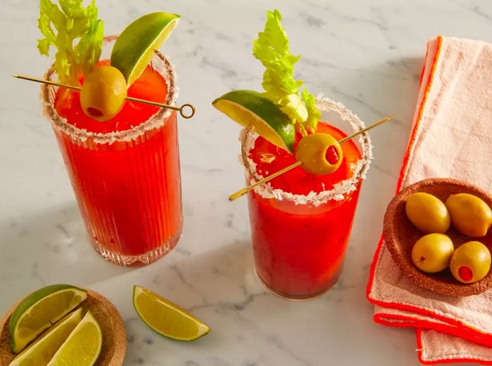 A high-angle view into two glasses of Bloody Mary, garnished with celery, lime, and olives.