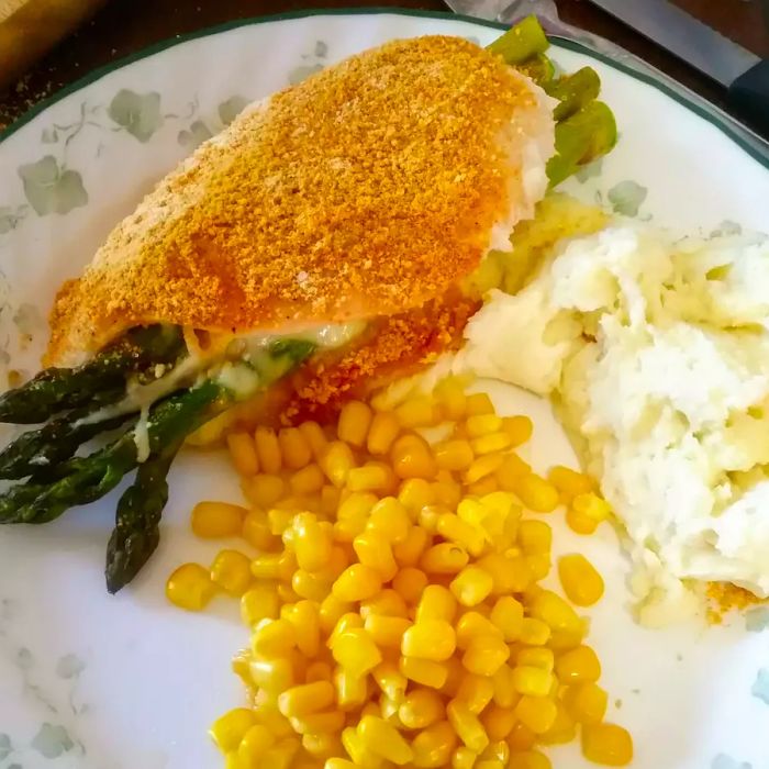 A close-up of Asparagus and Mozzarella-Stuffed Chicken Breasts, served with creamy mashed potatoes and corn on a white plate.