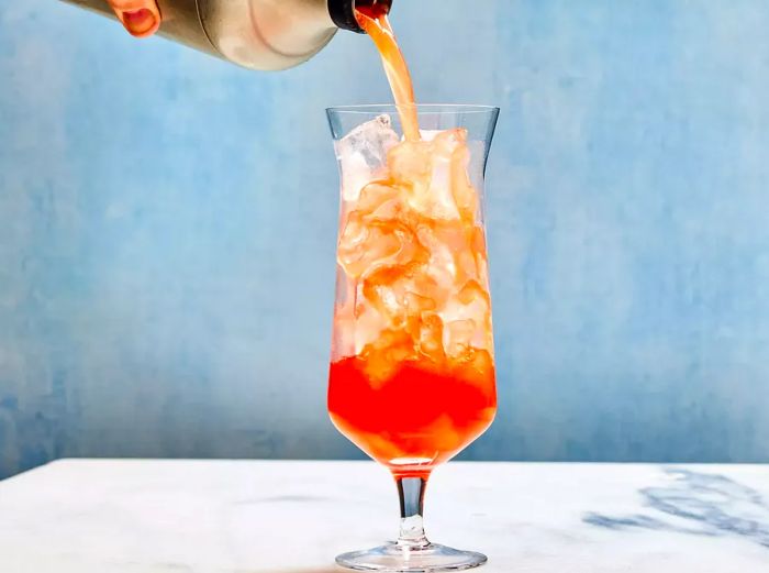 A cocktail shaker pouring the drink into a hurricane glass filled with ice