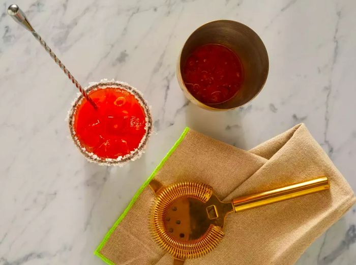 High-angle shot of a Bloody Mary in a glass next to a strainer and a shaker with leftover cocktail.