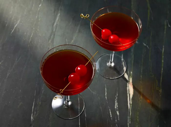 A close-up view of two coupe glasses filled with Manhattan cocktails, each garnished with cherries.