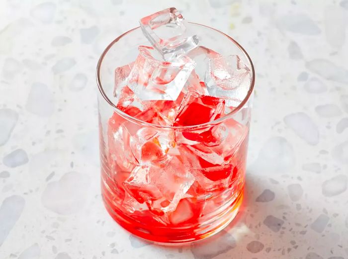 A high-angle shot of a rocks glass filled with ice and a splash of red grenadine syrup